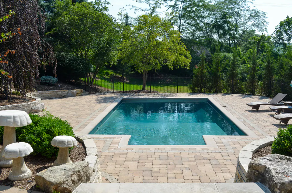 Long poolside view with rectangular pool, paver deck, umbrella, lounge seating, limestone wall, and wooded edge.